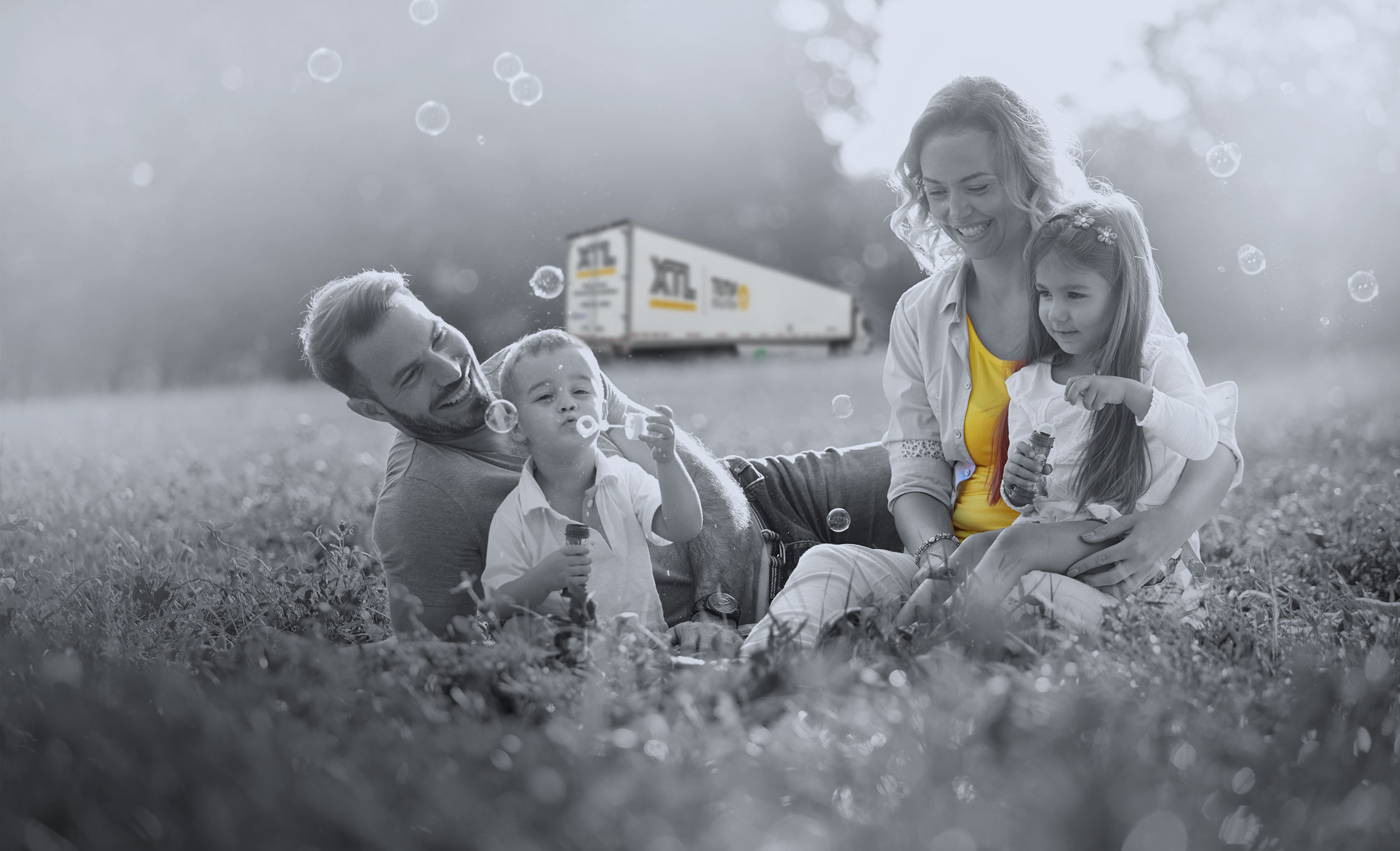 Family playing in the grass, blowing bubbles, with XTL transport truck in the background