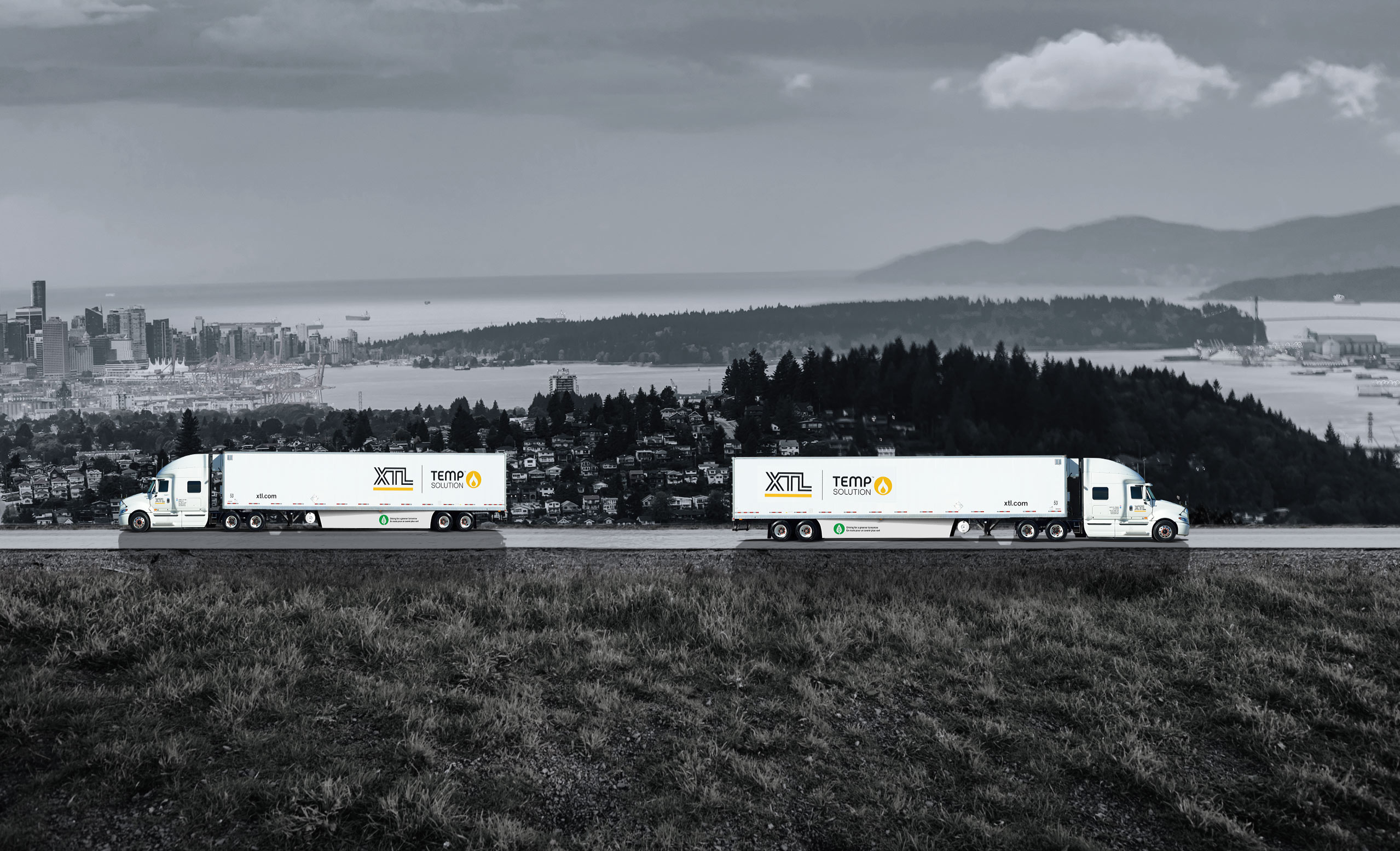 Two XTL transport trucks with trailers driving on the road with city of Vancouver in the background