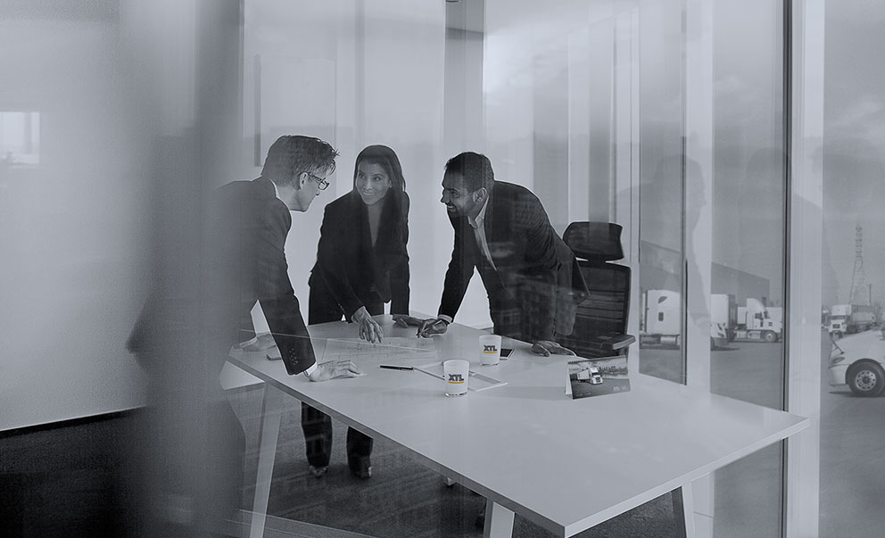 Trois personnes d’affaires debout autour d’une table de la salle du conseil de XTL avec camions à l’extérieur visibles par la fenêtre